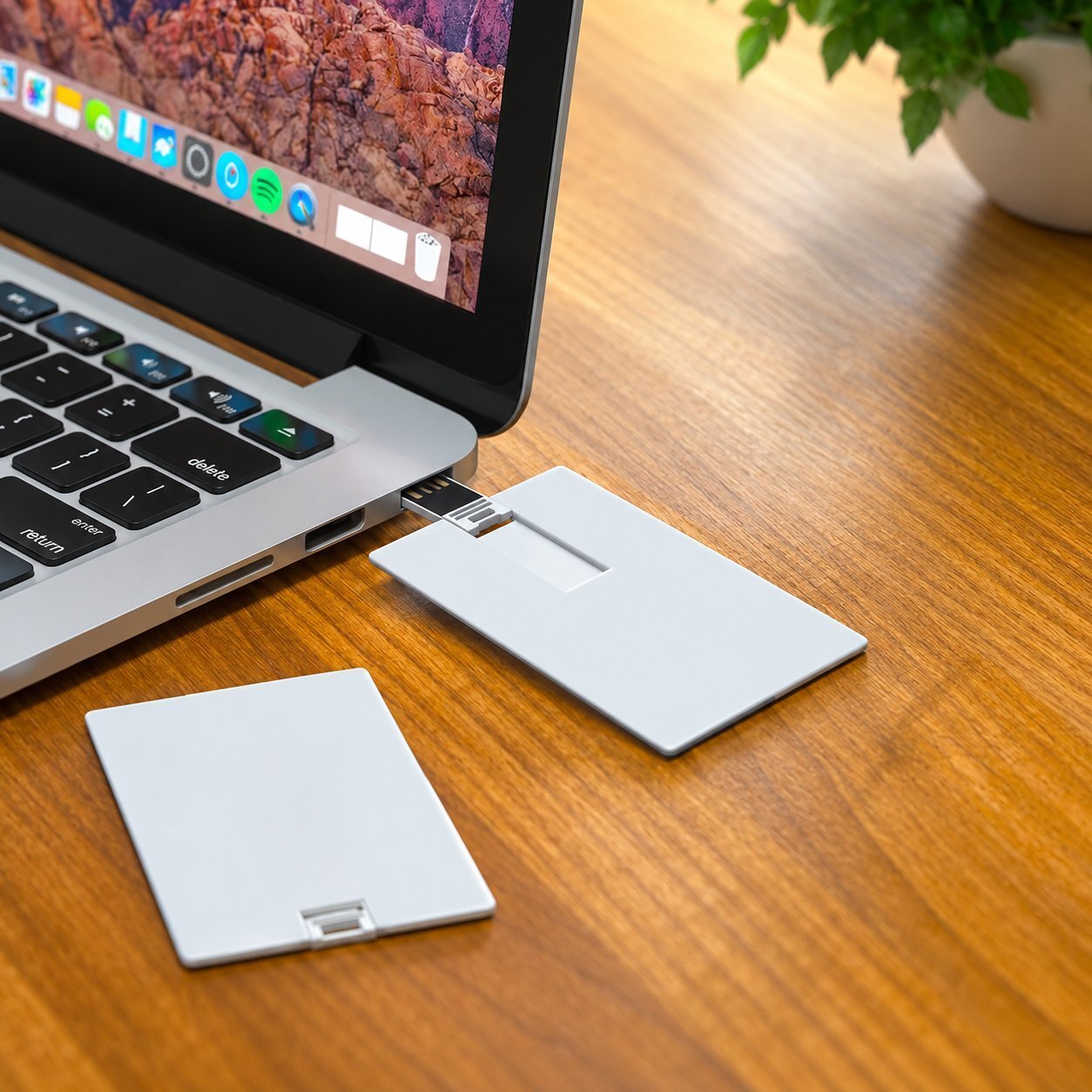 Two slim white plastic credit-card style USB drives on a wooden desk next to a laptop, one plugged into the USB port, showing the thin plastic card form factor used for printed marketing handouts.