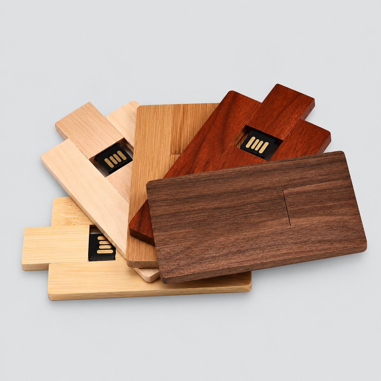 A stack of wooden card-shaped USB flash drives in light maple, bamboo, cherry, and dark walnut finishes, each with a sliding USB connector exposed at the top, illustrating the wooden USB casing option for branded corporate gifts.